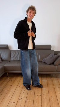 Jachetă din fleece neagră cu guler înalt, purtată peste un tricou alb, asortată cu blugi largi din denim albastru și pantofi negri lucioși.