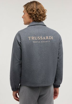 Homme aux cheveux bouclés portant une veste grise avec le texte "TRUSSARDI GENTLE SOCIETY" dans le dos, regardant vers la gauche sur un fond uni.