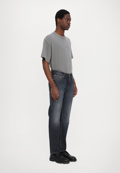 Grå t-shirt med kort ärm, mörkblå denimjeans, svarta snörskor; modellen står på sidan mot en vit bakgrund.
