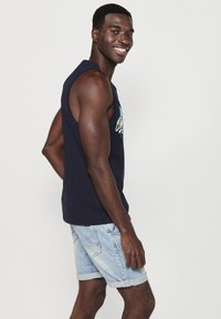 Tumedast tank top lahtise lõikega, mille ees on helesinise graafilise tekstiga print. Koos kergete rullitud äärtega denimpükstega.