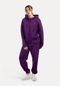 Sudadera con cremallera en color púrpura con grandes bolsillos y pantalones de chándal a juego. Hecha de tela suave, cuenta con puños elásticos y capucha. Zapatillas blancas.