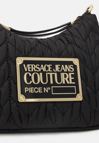 Borsa in tessuto trapuntato nero con una placca dorata che reca la scritta "Versace Jeans Couture Piece No" in lettere in rilievo.