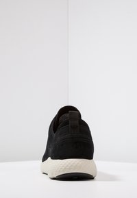 Svart mockaskor med en strukturerad vit sula. Har en dragflik vid hälen och en slät, ren design.