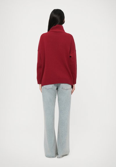 Pull en tricot rouge avec un col montant et des bords côtelés, associé à un jean large bleu clair. Design minimal, texture douce.