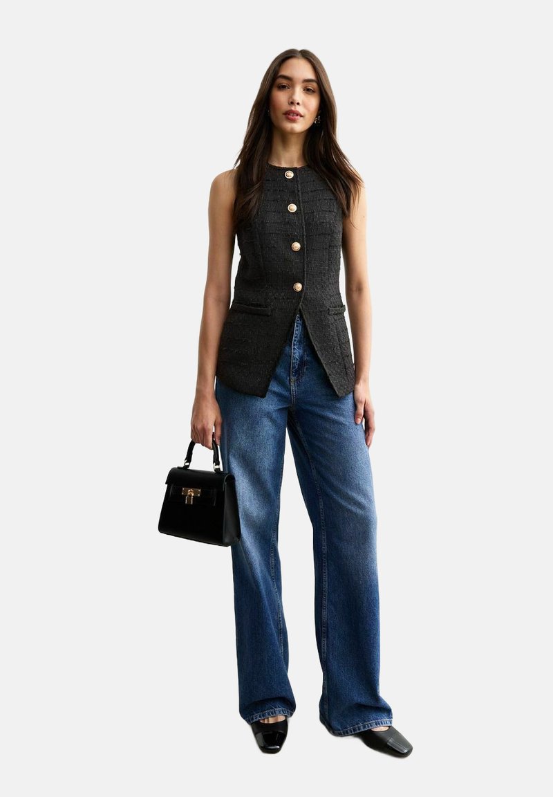 Graue Weste mit goldenen Knöpfen, kombiniert mit weiten blauen Jeans. Hält eine kleine schwarze Handtasche. Glatte Textur, maßgeschneiderte Passform und ohne Ärmel.