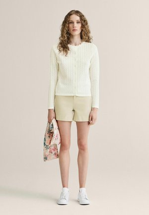 Kvinde med krøllet hår iført en hvid strikket cardigan, beige shorts, hvide sneakers, der holder et blomsterstof, stående mod en lys baggrund.