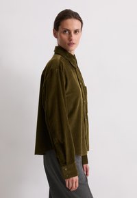 Chemise en velours côtelé vert olive avec une fermeture à boutons à l'avant, un col et une poche poitrinaire unique. Manches longues avec des poignets à boutons. Texture à côtes verticales.