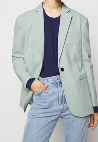 Ljusgrön blazer med texturerad yta, en svart knapp, och slagkragar, kombinerad med en marinblå topp och höga ljusblå jeans.