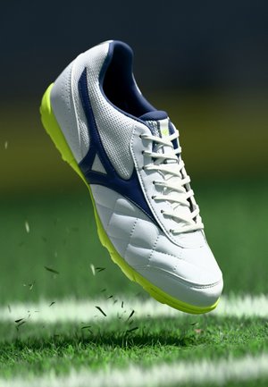 Chaussure de football blanche et marine avec semelle jaune néon en l'air au-dessus d'un terrain de pelouse verte avec ligne blanche.