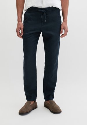 Homme portant un pantalon bleu marine à cordon et des chaussures en daim marron à enfiler, debout avec les mains relâchées le long du corps sur un fond blanc.