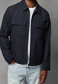 Giacca blu navy con zip frontale, due tasche sul petto e maniche lunghe. Indossata sopra una camicia bianca e abbinata a jeans azzurri chiari.