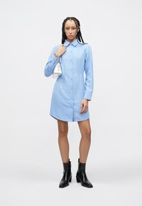 Lichtblauwe, lange mouwen overhemdjurk met knopen aan de voorkant, puntige kraag en zijnaden. Model draagt een kleine witte handtas en zwarte enkellaarzen.