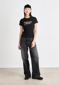 Svart bomullst-skjorte med "tommy jeans"-logo, kombinert med løstsittende mørkegrå denimjeans. Modellen har også på seg en svart caps og sko.