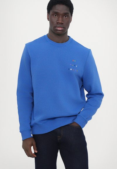Sweatshirt bleu en mélange de coton, avec un col rond, des poignets côtelés et un texte de logo coloré sur la partie gauche de la poitrine, porté avec un jean foncé.
