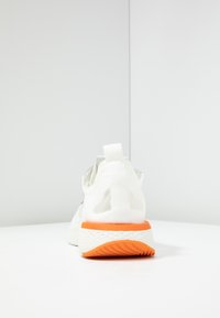White sneaker viewed from the back with orange sole edge and grip, textured midsole, and pull tab on heel against a plain white background.