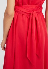 Section de robe rouge avec une ceinture nouée à la taille. Le tissu semble doux avec un design fluide et une texture lisse. Couleur unie simple sans motifs.
