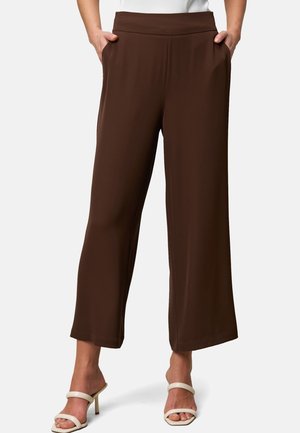 Pantalon marron à jambes larges et coupé porté avec des sandales blanches à talons, mains dans les poches, montré de la taille aux pieds.