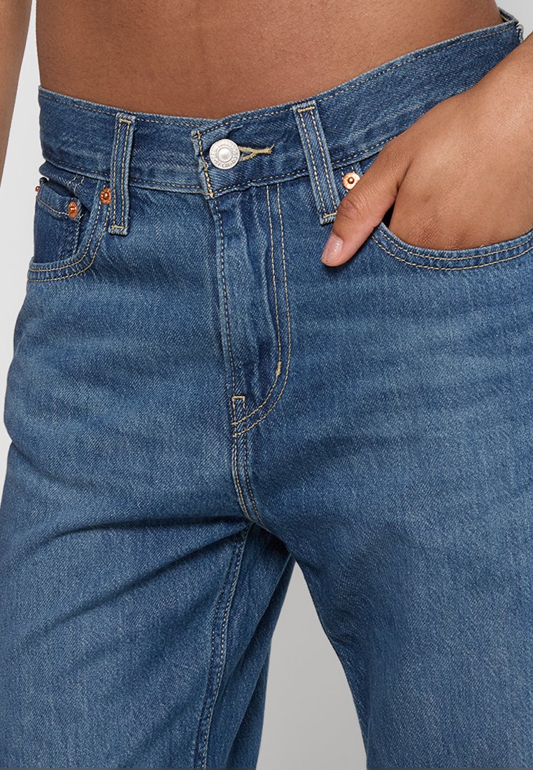 Gros plan sur une personne portant un jean en denim bleu avec une main placée dans la poche avant droite sur fond uni.