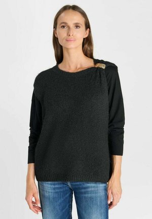 Donna che indossa un maglione senza maniche nero testurizzato con una spilla dorata sulla spalla sopra una camicia nera a maniche lunghe e jeans blu.