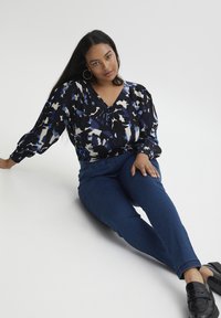 Blusa de manga larga con un patrón abstracto multicolor en azul, negro y blanco, combinada con jeans ajustados oscuros y zapatos negros tipo slip-on.