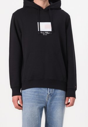 Mężczyzna w czarnej bluzie z kapturem Tommy Hilfiger z logo i napisem na piersi, w połączeniu z jasnoniebieskimi dżinsami, stojący na białym tle.