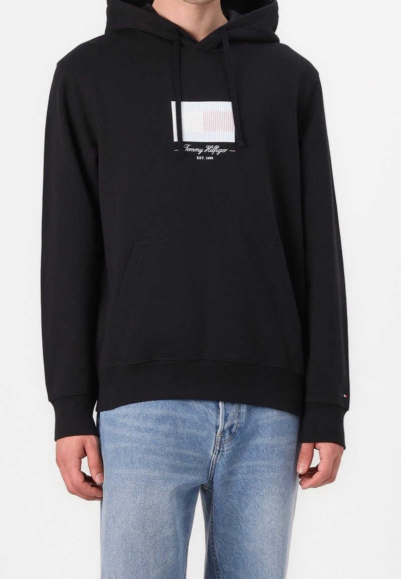 Homme portant un sweat à capuche noir Tommy Hilfiger avec logo et texte sur la poitrine, associé à un jean bleu clair, debout devant un fond blanc.