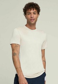Camiseta de manga corta color crema hecha de una tela suave, con un cuello redondo y un pequeño logo en el pecho. Textura lisa.