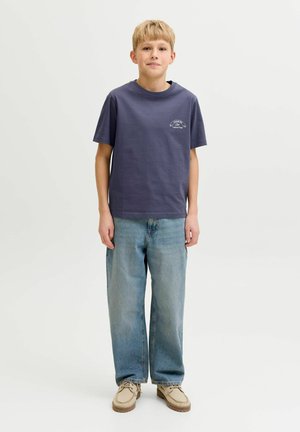 Blond chłopiec stoi, ubrany w ciemnoniebieską koszulkę z nadrukiem, luźne jasnoniebieskie dżinsy i beżowe buty na sznurowadła na tle gładkiej białej ściany.