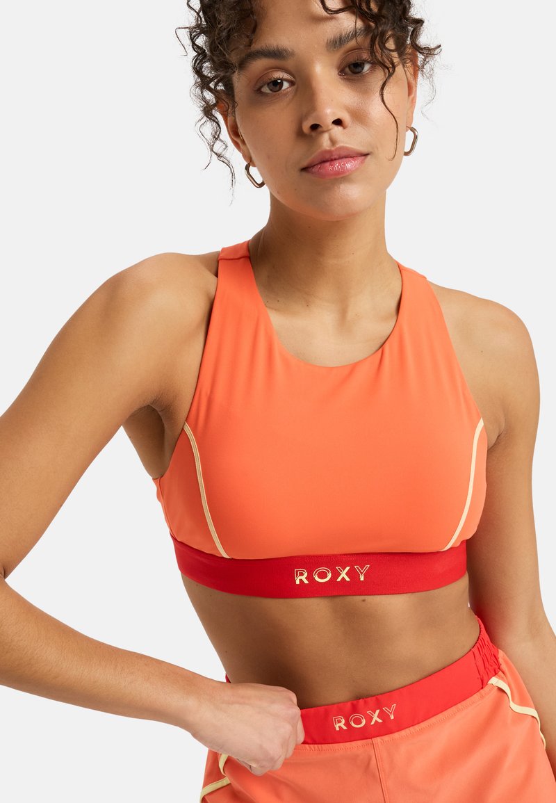 Orange idrotts crop top med rund halsringning, med ett rött band märkt "ROXY" och ljusa accenter längs sömmarna.