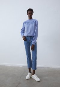 Felpa lilla con ricami floreali su spalle e maniche, abbinata a jeans a gamba dritta blu chiaro e sneakers bianche.
