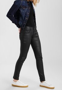 Svarta smala jeans i texturerad tyg, kombinerat med en mörk denimjacka och vita sneakers, vilket framhäver en snug silhuett.