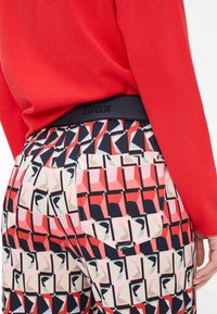 Colorful patterned pants featuring a geometric design in red, pink, and navy. Includes a black waistband with "Brax" logo and back pockets.