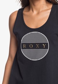 Zwart tanktop met een cirkelvormige afbeelding met witte strepen en het woord "ROXY" in gouden letters in het midden.