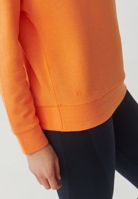 Felpa arancione con maniche lunghe e orlo a costine. Presenta un sottile dettaglio del logo vicino al fondo e una texture liscia e morbida. Indossata con leggings blu navy.