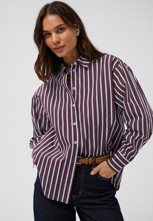 Femme aux cheveux bruns portant une chemise boutonnée à rayures verticales marron et bleu clair, un jean foncé et une ceinture marron, posant avec une main dans la poche.