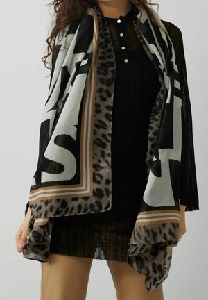 Femme portant un haut noir et un pantalon beige, tenant une grande écharpe aux motifs noir, blanc, marron et léopard drapée sur les épaules.