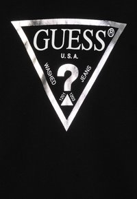 Tissu noir avec un logo triangulaire argenté comportant le texte "GUESS U.S.A." et "WASHED JEANS", ainsi qu'un symbole de point d'interrogation central.