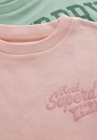Divas divas t-krekli: viens rozā ar izspiestu tekstu "Reāls Superdry", otrs zaļš ar tumši zaļu druku. Abiem ir apaļas apkakles.