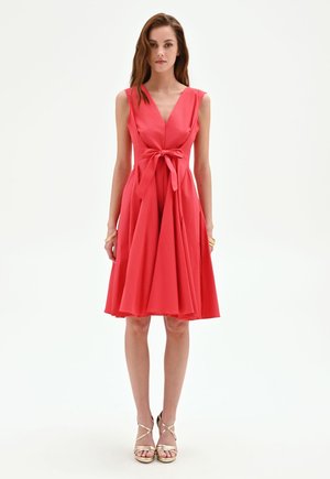 Femme portant une robe rouge sans manches avec un col en V et un nœud noué à la taille, associée à des sandales dorées à brides et talons hauts, debout sur fond blanc.