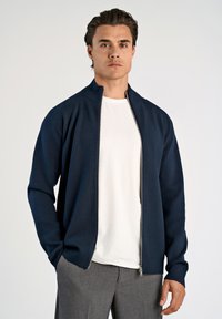 Navy vetoketjullinen takki, joka on valmistettu neuloksesta, korkea kaulus ja pitkät hihat, päällä valkoinen paita, jota täydentävät harmaat housut.