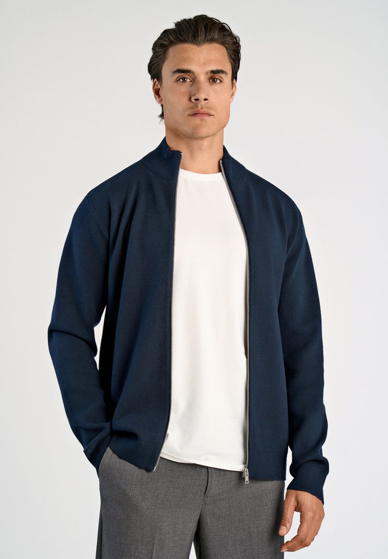 Granatowa kurtka zapinana na zamek, wykonana z dzianiny, z wysokim kołnierzem i długimi rękawami, noszona na białej koszuli, uzupełniona szarymi spodniami.