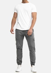 Man som bär en enkel vit t-shirt, grå cargobyxor med knäpaneler och elastiska muddar, samt vita sneakers mot en enfärgad ljus bakgrund.