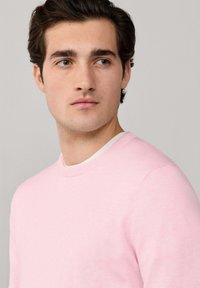 Maglione girocollo rosa chiaro realizzato in tessuto morbido. Presenta un logo discreto sul petto e una scollatura arrotondata. Texture liscia in tutta la superficie.