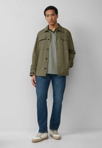 Giacca verde oliva con bottoni frontali e due tasche sul petto, maglietta grigia a scollo a V, jeans blu e sneakers beige con accenti bianchi.