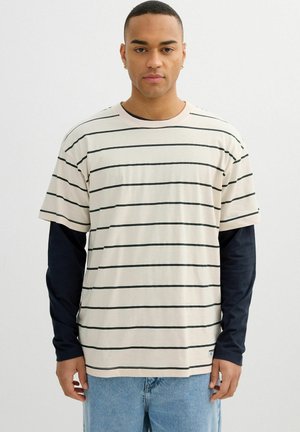 Hombre que lleva una camiseta beige de manga corta con rayas horizontales negras superpuesta sobre una camiseta negra de manga larga y vaqueros azul claro.