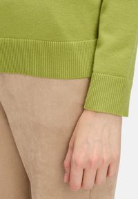 Zöld kötött pulóver bordázott mandzsettával, világos bézs velúr anyaggal párosítva, sima textúrával és testhez álló fazonnal az alján.