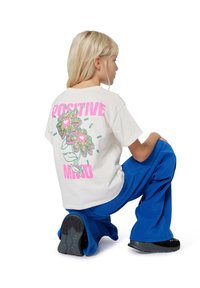 Witte katoenen t-shirt met een glitter bloemenprint in roze en groen en de tekst "POSITIVE MIND" op de achterkant; gecombineerd met blauwe wijde broek.