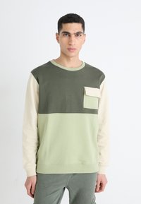 Sudadera color bloqueada en verde y beige con cuello redondo, que cuenta con un bolsillo frontal y puños acanalados. Tejido de algodón suave.