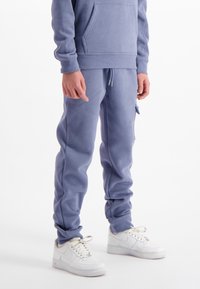 Persoon in blauwe joggingbroek met een zijzak en witte sneakers, staand tegen een plain witte achtergrond.