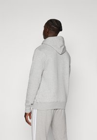 Hoodie grigio realizzato in morbido tessuto, con un grande cappuccio, polsini e orlo a coste. Indossato con pantaloni della tuta grigi abbinati con strisce bianche.